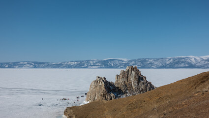 An unusual two-headed mountain, devoid of vegetation, rises above a frozen lake. Steep slopes, cracks in the rocks. Tiny silhouettes of people on the ice. A mountain range against the blue sky. Baikal