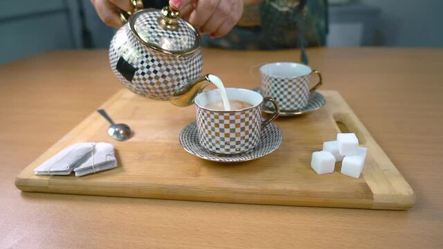Female pouring hot milk into a cup to make delicious tea at home - Indian chai. Tea-making ingredients tea bags  sugar cubes  and a spoon kept together on a wooden board in the kitchen