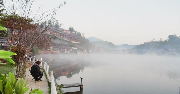Beauty in nature Morning view and tourist woman sitting relaxing at riverside with lake fog misty and Chinese style village hill on background at Ban Rak Thai village, Mae hong son, Thailand