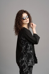 Photo of charming woman 20s with long curly hairstyle wearing trendy sunglasses, showing emotions, with fashion look over gray background
