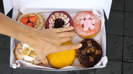Top view of vegan donuts with different frostings and fillings. A hand picking yellow mango one from the carton box