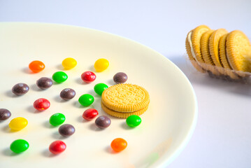 Multicolored, multiflavored candies in a white plate and cream sandwich cookies on a white background