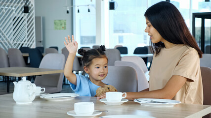 Mother with kid girl in restaurant. Toddler child with space buns raises hand with smile sitting by Asian mum at table with white tea cups in local cafe close view