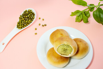 Close-up of broken mung bean cake on white plate, mung beans in a spoon, green plant, pink background, top view