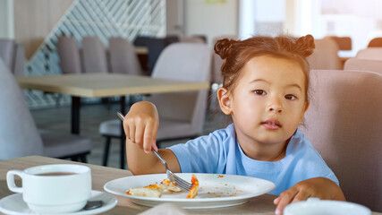 Kid eats pizza. Cute Asian toddler girl in blue t-shirt eats delicious snack with fork sitting on grey restaurant chair at table close view