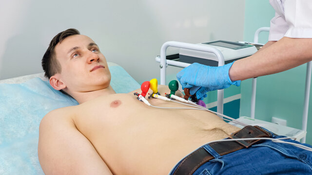 Nurse in gloves puts electrodes on bare chest of young man patient lying on couch to do electrocardiogram in light hospital office close view