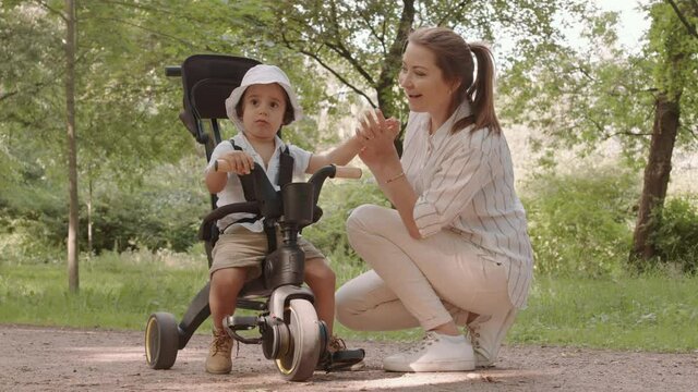 Young affectionate mother and her cute toddler boy sitting in tricycle stroller spending time outdoors in park on warm sunny day