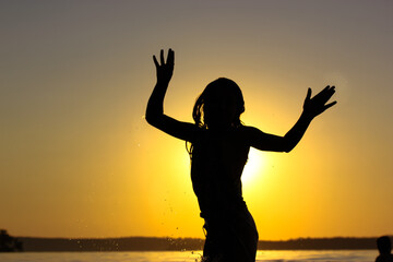 Fototapeta premium Contrast silhouette of a child teenage girl frolicking in a water body, river, lake, sea, ocean against a yellow sunny sunset sky. Place for text. 