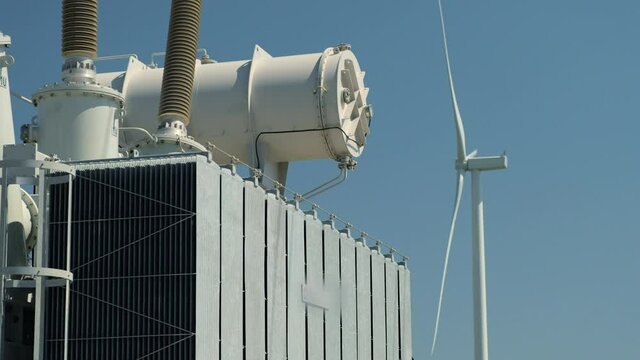 High potential power transformer receives electricity generated by wind turbine against operating windmill under blue sky on sunny day