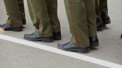 Soldiers in green trousers and black shoes march in place on the parade ground. Officers in uniform stride along the white line.