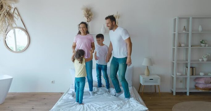 4K video footage with happy middle aged parents jumping on the bed with their adorable children, school boy and younger girl. Full length carefree two generations family