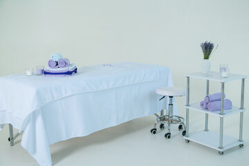 Massage couch covered with white terry sheet. Chair and table with towels, candles and a vase with a bouquet of lavender. All on a white background.