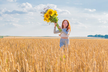 Young happy red-haired girl with a large bouquet of sunflowers and wildflowers in her hands runs across a yellow wheat field against a blue cloudy sky