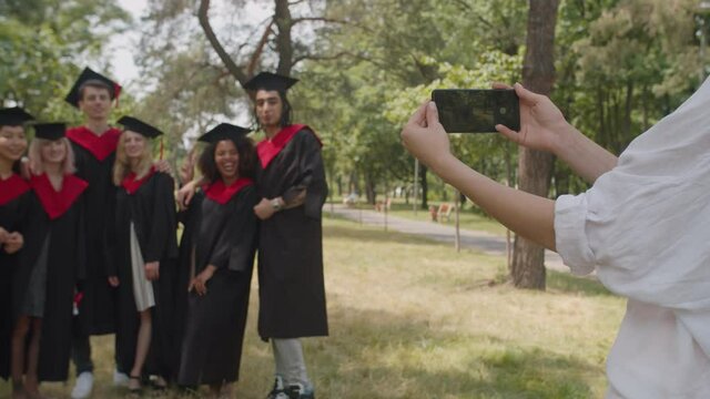 Close-up of female hands taking photo of blurry carefree diverse multiracial graduates in graduation gowns and mortarboards on smart phone outdoors after graduation ceremony. Focus on phone screen.