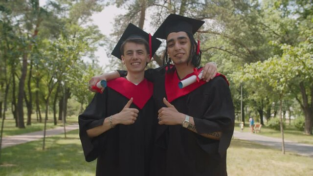 Portrait of successful proud handsome multiracial male graduates with diploma in graduation gowns and mortarboards embracing, showing thumbs up, expressing cheerful mood and happiness outdoors.