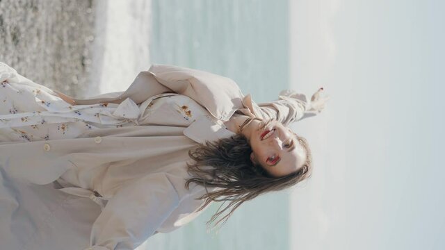 Young woman in trench coat walking on beach. Vertical video. 4k footage