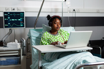 Sick young patient with headphones using laptop in hospital ward. African american teenager with...
