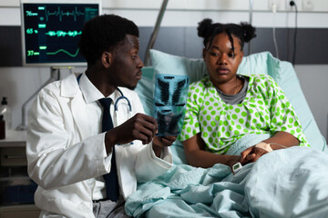 Obraz premium Afro specialist analyzing x ray results with young patient laying in hospital ward bed at clinic. African american doctor showing radiography scan to girl with disease, learning diagnosis