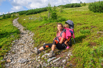 Naklejka na ściany i meble A mother with her sleeping daughter in a carrier backpack is resting on the grass before climbing a stone path in the Carpathian Gorgan. Ukraine