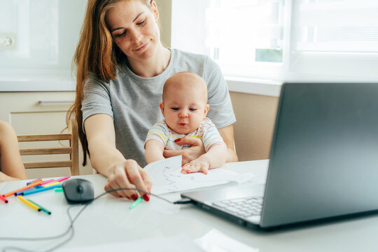 A Young Happy Smiling Mother With A Baby In Her Arms Sits At The Table And Works On A Laptop And With Documents.