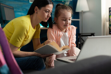 Fototapeta premium Mother and daughter doing homework for online school class with teacher sitting at home desk. Adult parent giving education knowledge to little child preparing for lesson lecture on laptop