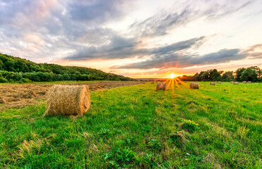 Scenic view at sunset or sunrise in green shiny field with hay stacks, bright cloudy sky, golden sun rays, summer valley landscape