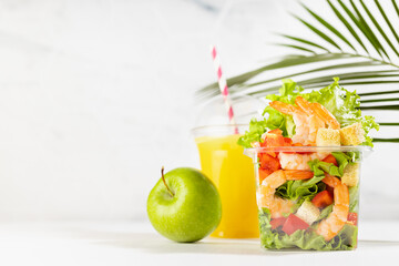Healthy summer tropical take away food set of orange juice, green apple, salad with shrimps, lettuce, red bell pepper, croutons in plastic box on white wood table, marble tile wall with palm leaf.