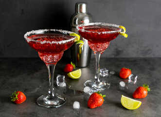 Two glasses of strawberry margarita with ice, mint and lime peel. Shaker, strawberry berries and lime slices in the background