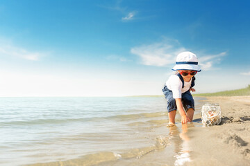 Kid play on the beach on a hot sunny day. Little girl walks barefoot along the sandy seashore and collects shells in a jar.