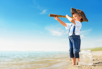 Kid play on the beach on a hot sunny day. Little girl dressed as a pirate stands barefoot on the sandy seashore with a telescope. Child with a toy fish dreams of travel and adventure. - obrazy, fototapety, plakaty