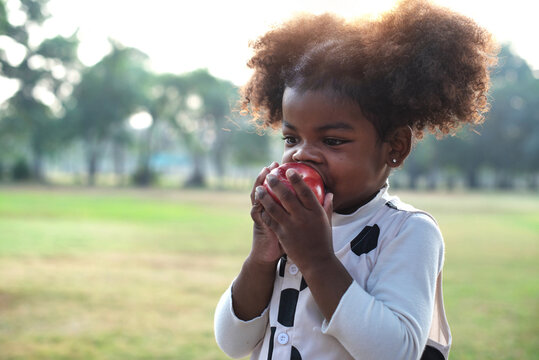 Portrait of happy little dark skinned girl eating red apple outdoor garden