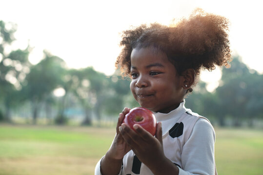 Portrait of happy little dark skinned girl eating red apple outdoor garden