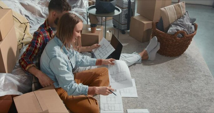 High angle young man and woman work with housing apartment plan sheets sitting among removal boxes after moving in.