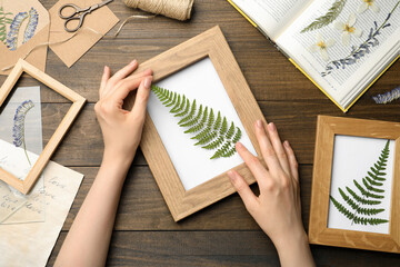 Woman making beautiful herbarium with pressed dried fern leaf at wooden table, top view.