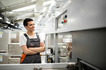 Portrait of short haired female industrial worker in working uniform standing in factory by manufacturing machine.