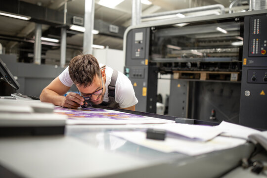 Professional print worker checking imprint quality and color matching with magnifying glass in printing house.