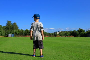 Obraz premium Self confident kid standing up at a green lawn. Gray shirt and shorts with a blue cap. Back towards camera. Nice summer day outside. Stockholm, Sweden, Europe.