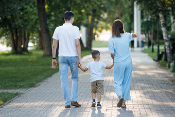 Family - mom, dad and son are walking along the pavement in the city summer park. Back view.