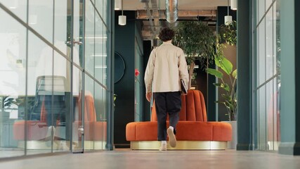 Full length shot of casually dressed young man walking along corridor of modern open plan office before sitting on orange circular sofa and taking out laptop to work in slow motion