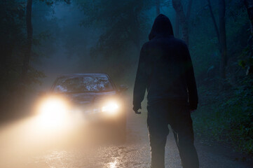 A spooky hooded figure silhouetted against a cars headlights. On a scary forest road at night.