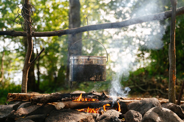 A tourist soot-covered bowler hat hangs over a smoking campfire at a forest camp in summer. Camping cooking.