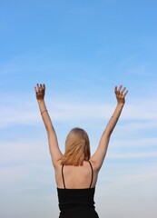 A young caucasian woman with her hands up in a black T-shirt against a blue sky. The concept of freedom, happiness, success, health. Back view