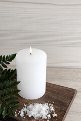 Burning candle, sea salt and green leaves on light beige wooden table