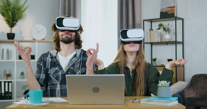 Attractive happy loving modern young couple sitting at the table at home and meditating under relaxed peaceful vision in augmented reality headsets,modern technology concept
