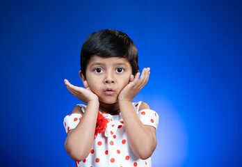 Cute surprised girl kid saying wow by closing mouth with hands and looking at camera on blue background - concept of crazy deal or offers.