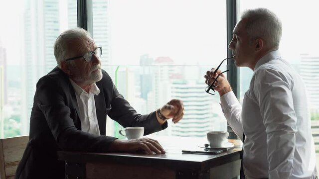 two senior businessman take a break drinking coffee and discussing business strategy during break on table in coffee shop at modern restaurant on a tall building cityscape background