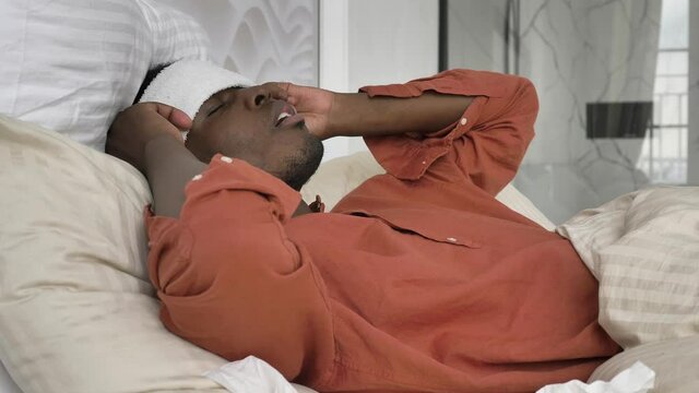 Upset African-American man with patch on head pills and thermometer suffers from cold lying on large bed at home close view