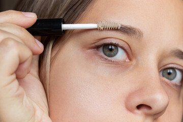 Close-up of the face of a young caucasian woman applying an eyebrow care gel with a brush. Firming agents for eyebrow growth - obrazy, fototapety, plakaty