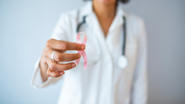 Doctor with pink ribbon and stethoscope on white background. Breast cancer awareness. Close up background image of unrecognizable female doctor holding pink ribbon as symbol of breast cancer awareness