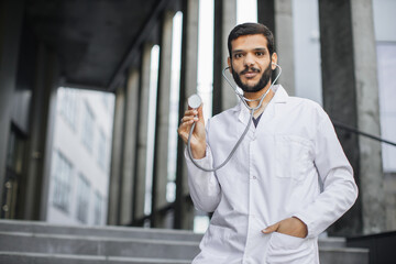 Waist up portrait of smiling pleasant male Indian doctor or medical student in white coat, standing outside hospital or university, demonstrating his stethoscope to camera. Panorama, copy space